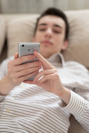 Portrait of teenager young man lying on sofa at home, holding and using smart phone. Focus on the phoneの写真素材