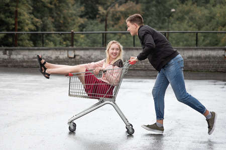Young couple having fun on shopping trolley. Happy man pushing shopping cart with his girfriend...