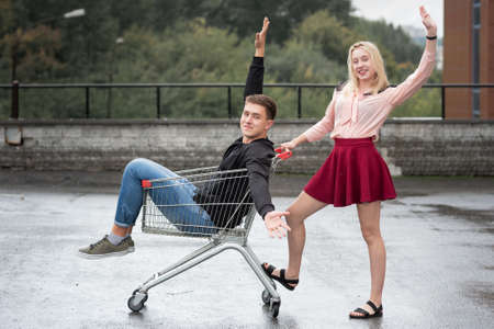 Young couple having fun on shopping trolley. Happy woman pushing shopping cart with her boyfriend inside on in the streetの写真素材