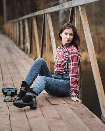 Young woman sitting whith ukulele guitar on bridge in autumn forestの写真素材
