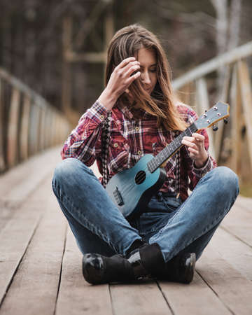 Young woman sitting and playing ukulele guitar on the bridge in autumn forestの写真素材