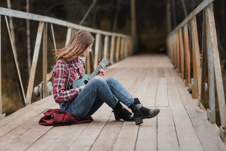 Young woman sitting and playing ukulele guitar on the bridge in autumn forestの写真素材