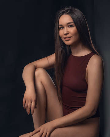 Studio photo of pretty brunette woman sitting on cube and hugging her leg with hand on black background. Model looking at cameraの写真素材