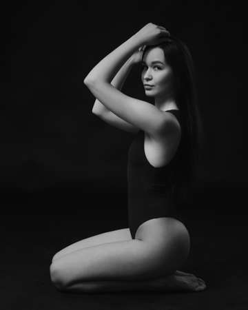 Studio photo of pretty brunette woman wearing in a black bodysuit sitting on the studio floor on black background. Model looking at camera. Black and whiteの写真素材