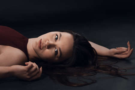 Studio photo of pretty brunette woman wearing in a black bodysuit sitting on the studio floor on black background. Model looking at cameraの写真素材