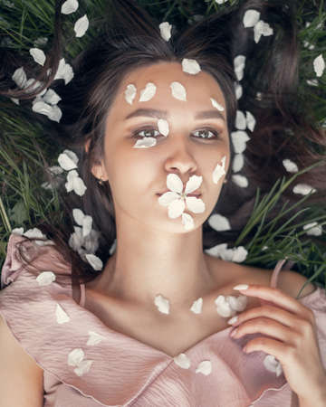 Girl with apple tree flower in lips and petals on face lies on the grass in spring