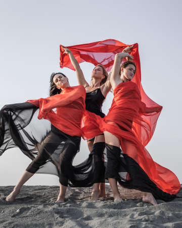 Three girls in black and red flowing fabric romantic pose and dance on sand duneの写真素材