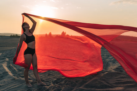 Elegant girl performs dance moves on the sand holding up long red fabric against sunset.の写真素材