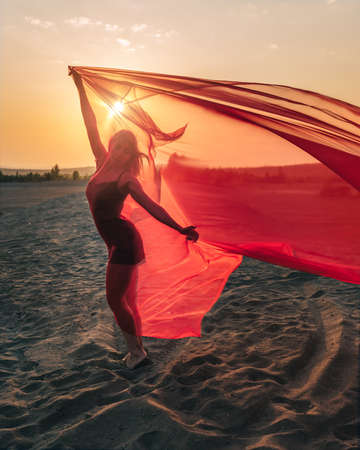 Elegant girl performs dance moves on the sand holding up long red fabric against sunset.の写真素材