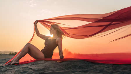 Elegant girl sitting on the sand holding up long red fabric against sunset.