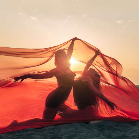 Elegant girl perform dance moves on sand holding up long red fabric against sunset.の写真素材