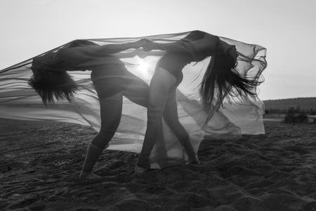 Elegant girl perform dance moves on sand holding up long red fabric against sunset. black and whiteの写真素材