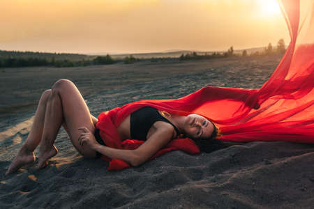 Elegant girl is on the sand holding up long red fabric against sunset.の写真素材