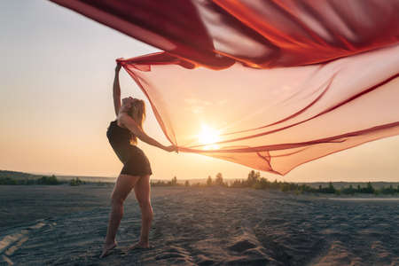 Elegant girl performs dance moves on the sand holding up long red fabric against sunset.の写真素材