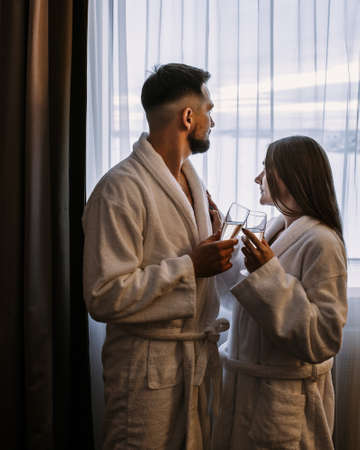 Couple wearing white bathrobes in hotel standing near window with glasses of champagneの写真素材