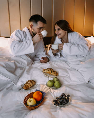 Happy young couple sitting in bed in morning and having breakfastの写真素材