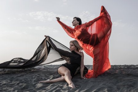 Two girls in black and red flowing fabric romantic pose and dance on sand duneの写真素材