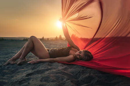 Elegant girl is on the sand holding up long red fabric against sunset.の写真素材
