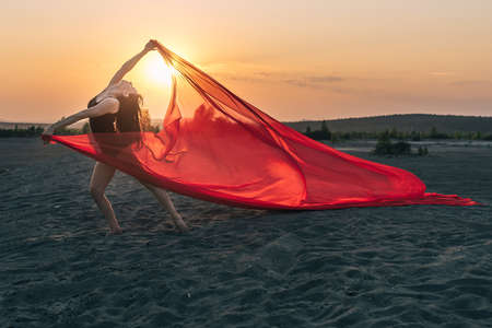 Elegant girl performs dance moves on the sand holding up long red fabric against sunset.の写真素材