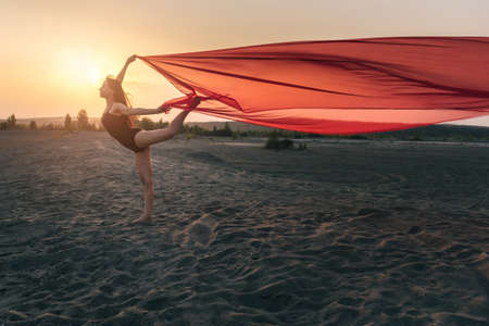 Elegant girl performs dance moves on the sand holding up long red fabric against sunset.の写真素材