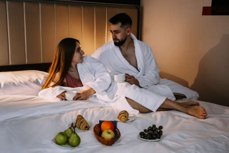Happy young couple sitting in bed in the morning and having breakfast in hotelの写真素材