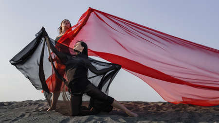 Two girls in black and red flowing fabric pose and dance on sand duneの写真素材
