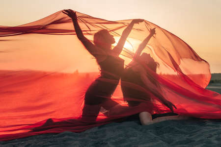Elegant girl perform dance moves on sand holding up long red fabric against sunset.の写真素材