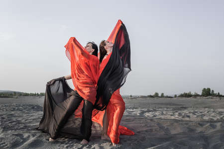Two girls in black and red flowing fabric romantic pose and dance on sand duneの写真素材