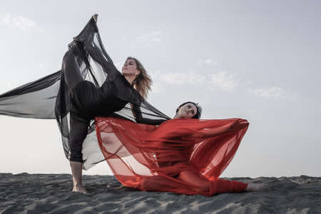 Two girls in black and red flowing fabric romantic pose and dance on sand duneの写真素材