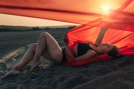 Elegant girl is on the sand holding up long red fabric against sunset.の写真素材