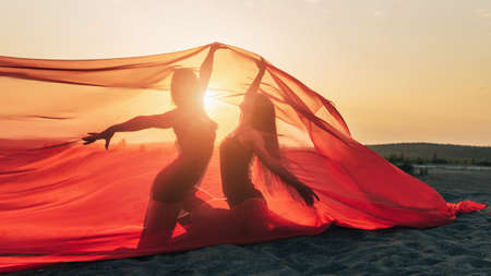 Elegant girl perform dance moves on sand holding up long red fabric against sunset.の写真素材