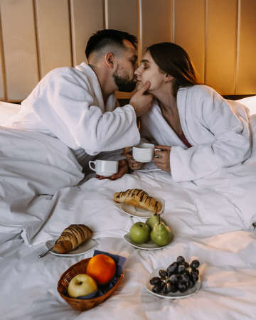 Happy young couple sitting in bed in the morning and having breakfast in hotelの写真素材