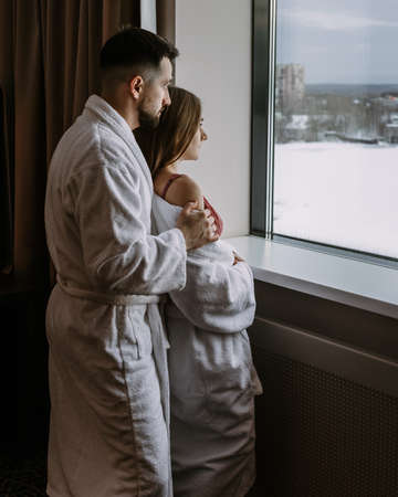 Couple wearing white bathrobes in hotel standing near windowの写真素材