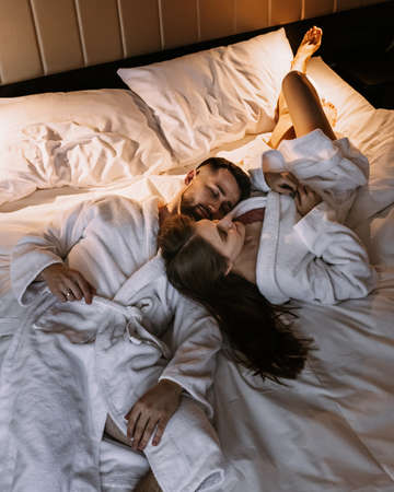 Young sexy loving couple in bathrobes relaxing on bed at hotelの写真素材