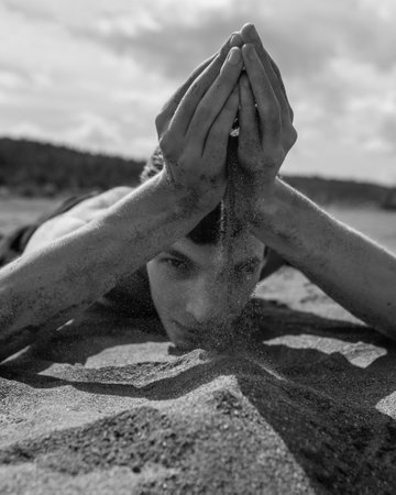 Young Man lying on his stomach and pouring sand through hands in the desert. Selective focus on hand in foreground.の写真素材