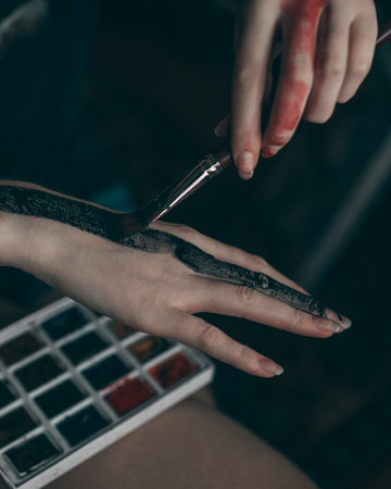 Young redhead woman artist applies red and black watercolor paint to her hand, with brushの写真素材
