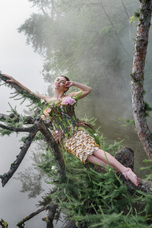 Blooming gorgeous lady in dress of flowers lying on tree over water in the morning fog. forest fairyの写真素材