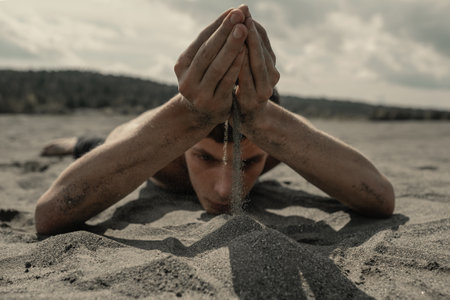 Young Man lying on his stomach and pouring sand through hands in the desert. Selective focus on hand in foreground.の写真素材