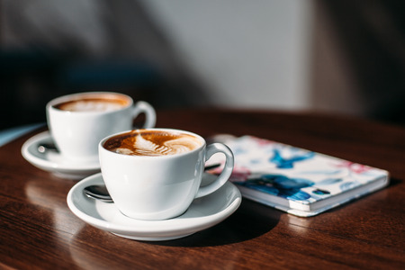 Two cups of cappuccino with latte art on wooden table. Beautiful foam, white ceramic cups, colourful notebookの写真素材