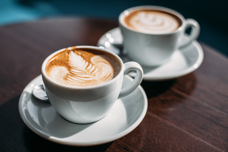 Two cups of cappuccino with latte art on wooden table. Beautiful foam, white ceramic cups.の写真素材
