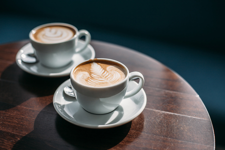 Two cups of cappuccino with latte art on wooden table. Beautiful foam, white ceramic cups.の写真素材
