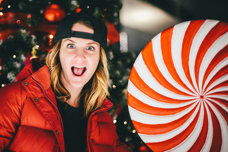 Trendy charming girl in red jacket wearing stylish cap with gigantic lollipop with Christmas tree on the backgroundの写真素材