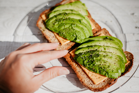 Whole grain toasts with avocado, cheese and herbs. A useful delicious breakfast. Atmospheric photo with hand, top view.の写真素材