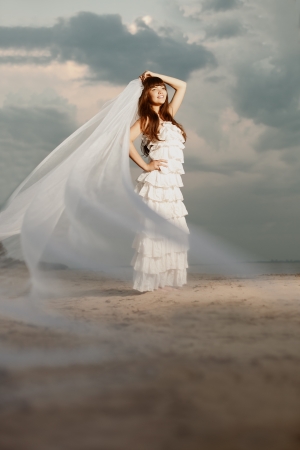 Beautiful bride in a long dress with a long veil on the beach at sunsetの写真素材