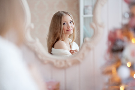 Beautiful young woman in white near the Christmas tree. Beautiful girl celebrates Christmas near the mirrorの写真素材