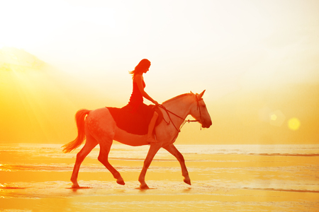 Woman and horse on the background of sky and water. Girl model on horseback on the beach by the sea at sunset, backlit in sunshine. A positive summer time scene.の写真素材