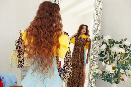 Girl trying on a dress in front of a mirror. Woman looks at purchases, fashionable clothes. Model with a leopard suit. Lady is going on a date, meetingの写真素材