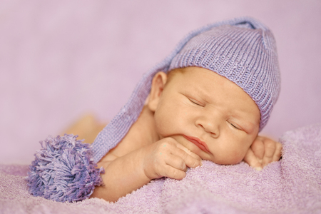 Little newborn baby in a funny hat sleeping in white blanket, lying on bed.の写真素材