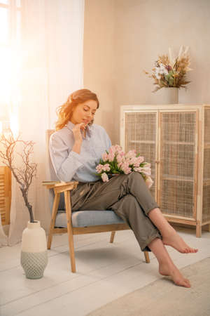 Young woman chilling at home. Girl relaxes and reads a book in the rays of the cozy warm sun. Sitting on retro chair near the window in light her living room in house with flowersの写真素材