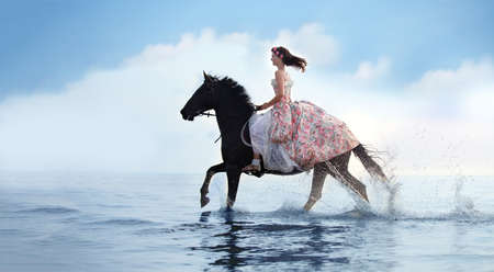 Beautiful girl in a long dress rides a black horse in the sea.の写真素材